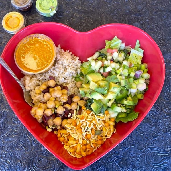 A heart-shaped bowl from Eats Meets West Bowls filled with rice, beans, salad, crunchy toppings, and a side of sauce.