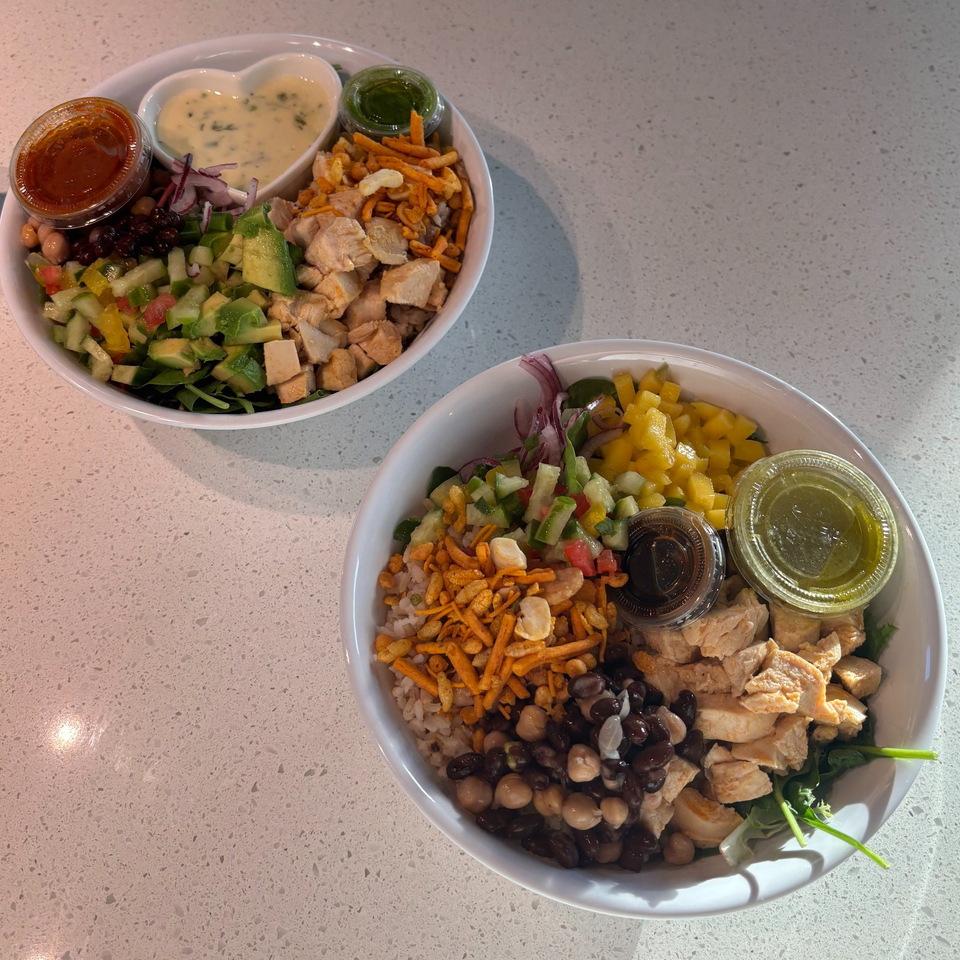 Two colorful grain bowls with chicken, beans, vegetables, avocado, crunchy toppings, and small cups of sauces on a countertop.