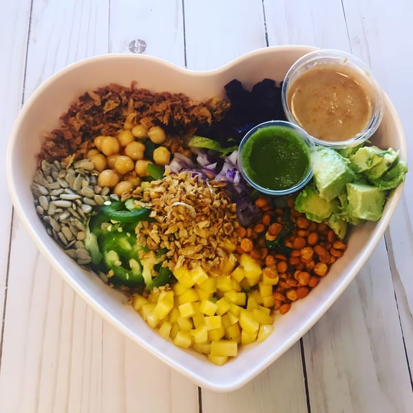 Heart-shaped bowl filled with chickpeas, mango, avocado, seeds, and two sauces on a white wooden table.